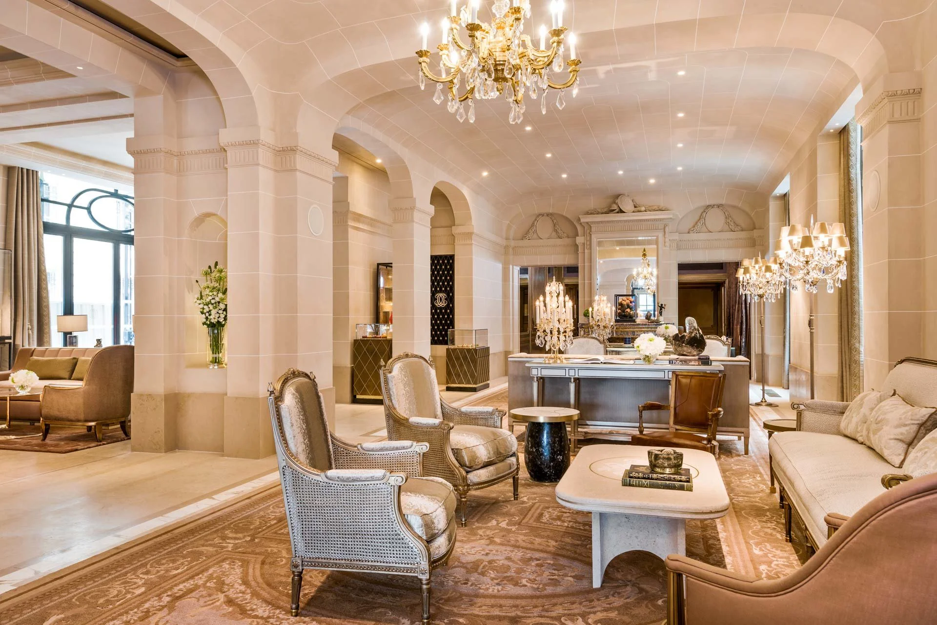 Salon élégant avec mobilier vintage, chandeliers en cristal, murs en pierre claire, grandes fenêtres avec rideaux, décor sophistiqué et ambiance chaleureuse.