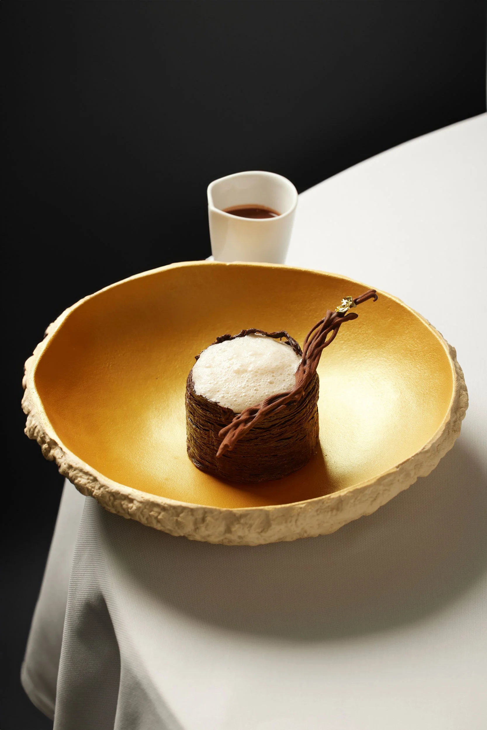 Un dessert au chocolat avec une mousse blanche, présenté dans un bol ovale doré avec un bord extérieur texturé. Une sauce ou une boisson est servie dans une petite tasse blanche derrière le dessert.