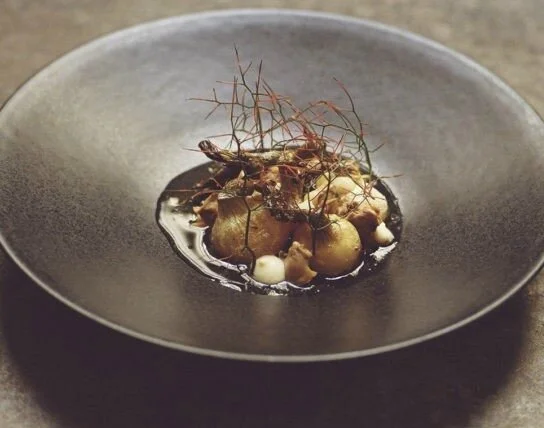 Assiette noire avec un plat gourmet comprenant des éléments végétaux, des petites boules blanches et une sauce noire.