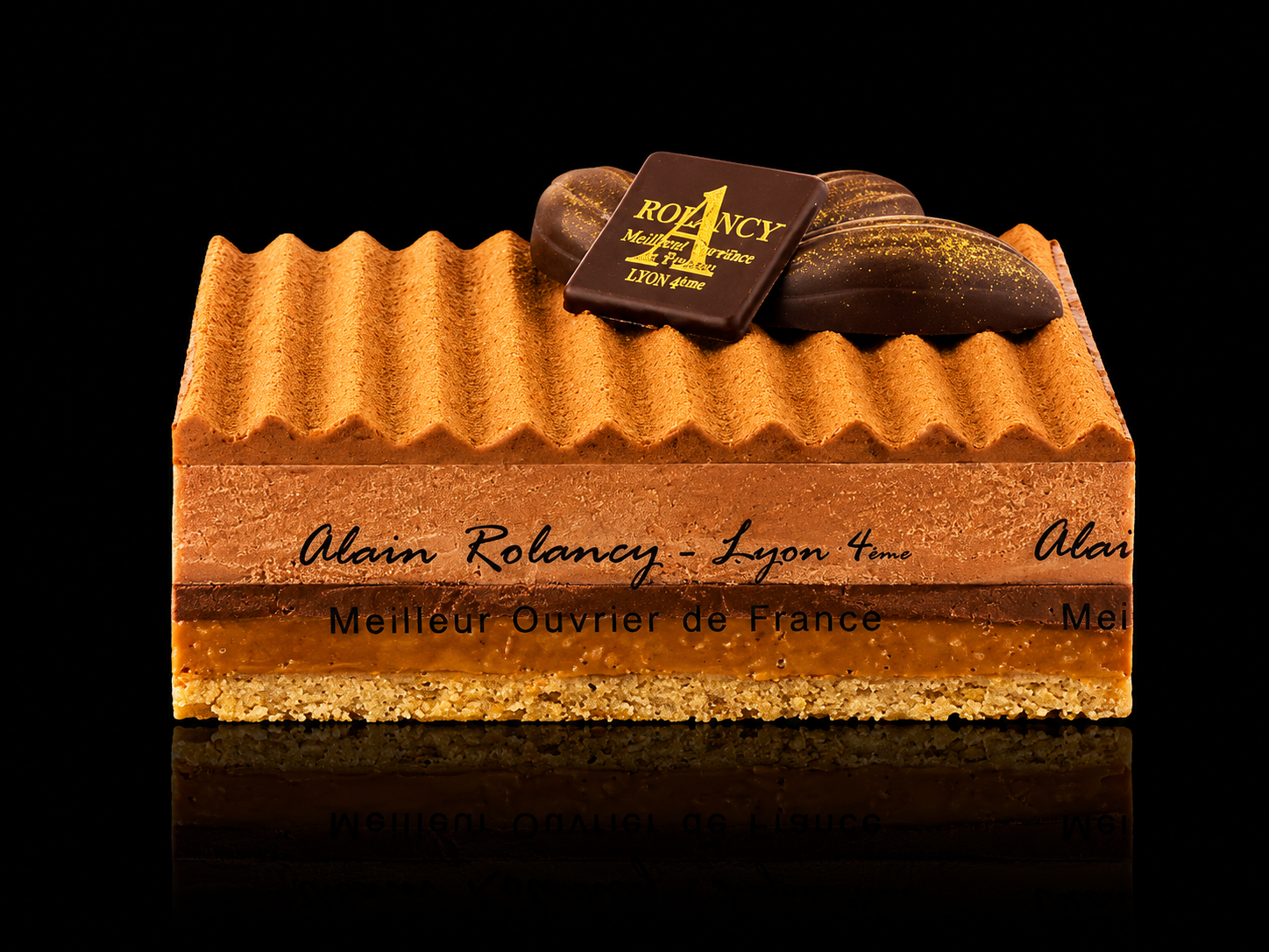 Gâteau décoratif au chocolat avec un dessus en forme de rayures, décoré d'un carré de chocolat portant l'inscription 'ROLANCY', sur fond noir.