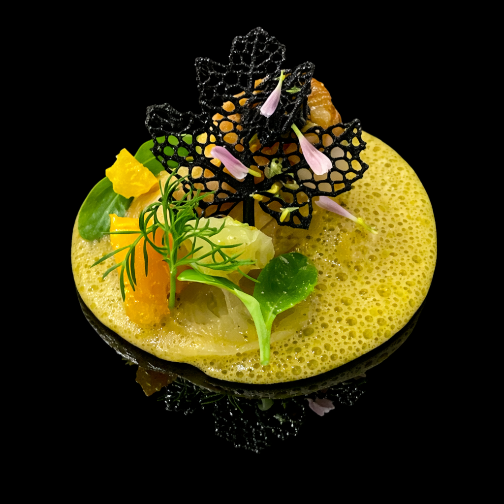 Assiette de cuisine gastronomique avec une mousse jaune, garnie de fleurs comestibles, de feuilles vertes et de déco noire en forme de lichen