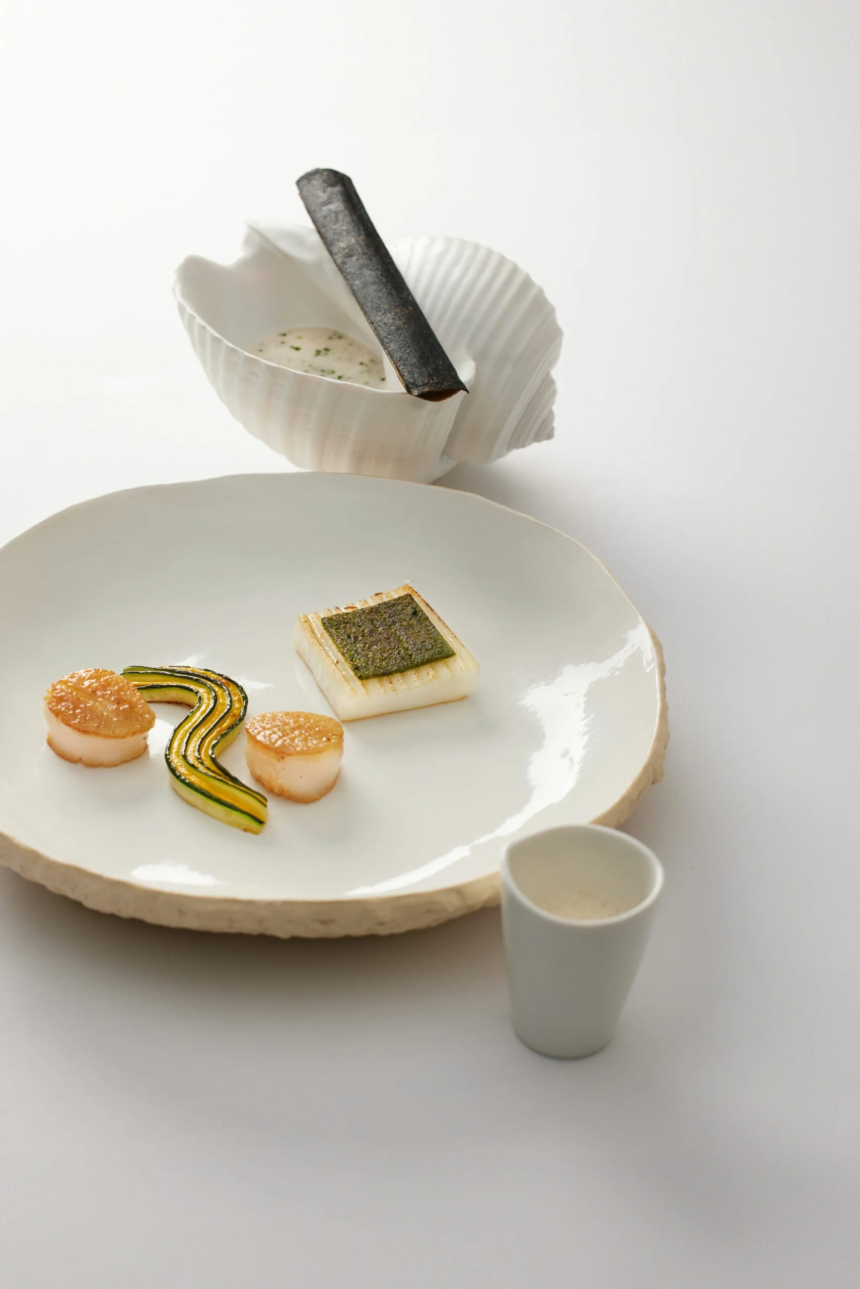 Assiette de cuisine créative avec divers éléments, servie sur une surface blanche, accompagnée d'une coquille avec une sauce et un bâtonnet carbonisé, et un petit verre