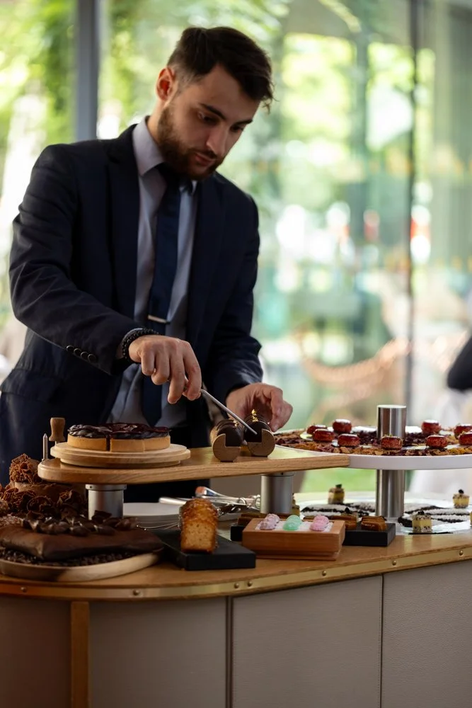 Un homme en costume ajuste ou coupe un gâteau lors d'une exposition ou d'une vente de pâtisseries dans une salle éclairée par la lumière naturelle grâce à de grandes fenêtres. La table est garnie de divers desserts, notamment des gâteaux et des pâtisseries.