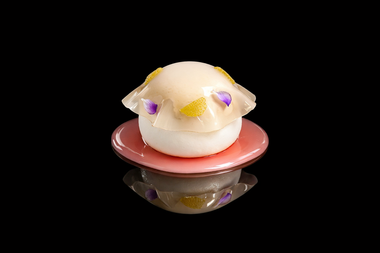Un dessert sophistiqué avec une boule de glace ou sorbet blanc, décoré de fines lamelles de gelée transparente, de morceaux de citron confit et de petites fleurs violettes, présenté sur une assiette rose sur fond noir.