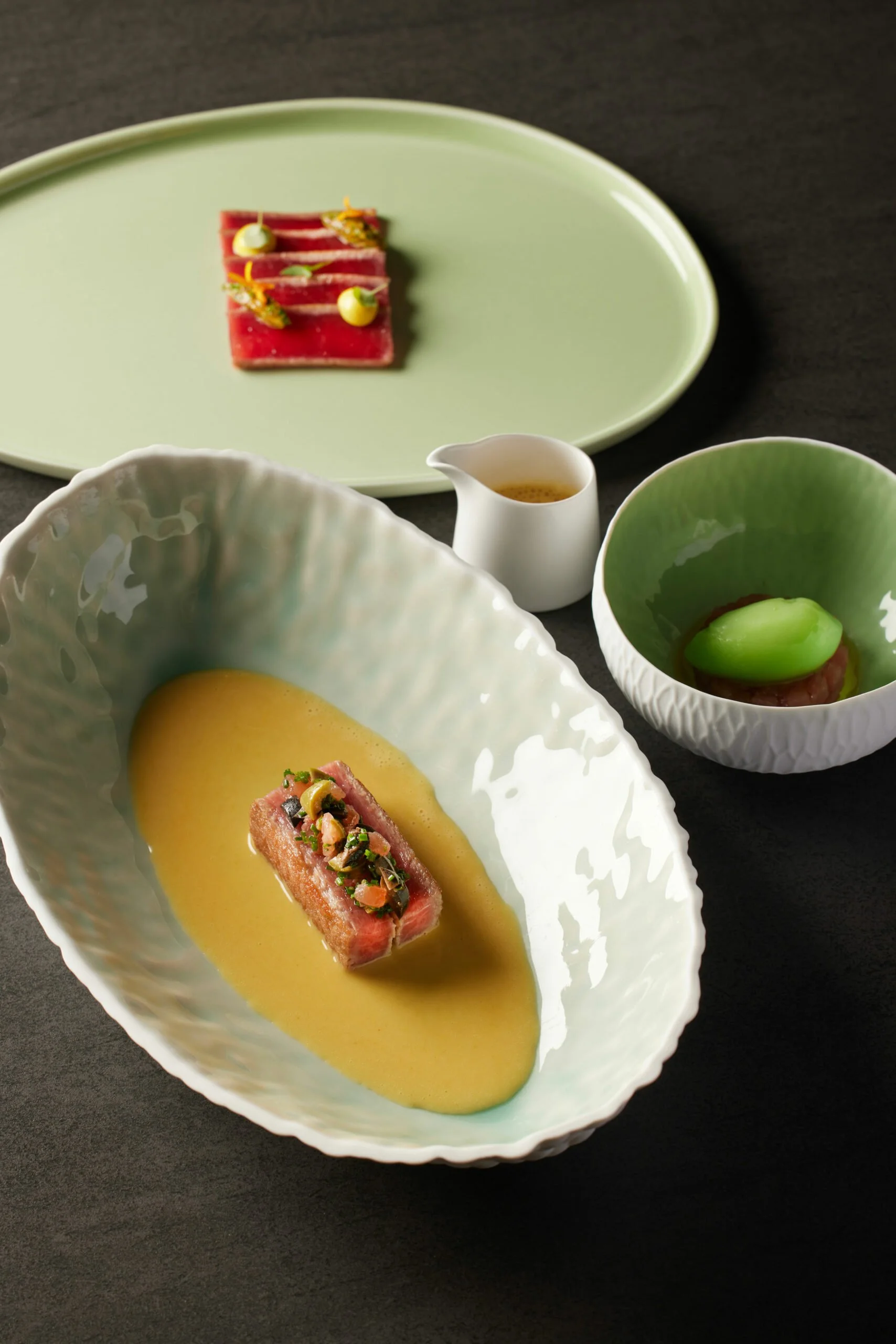 Assiette principale avec un morceau de steak servi dans une sauce jaune, garnie d'herbes et de légumes. Un petit bol à côté contiendrait probablement une sauce ou un condiment. Deux autres plats en arrière-plan avec des portions de viande ou de poisson décorées et une petite bouteille de sauce ou vinaigrette.