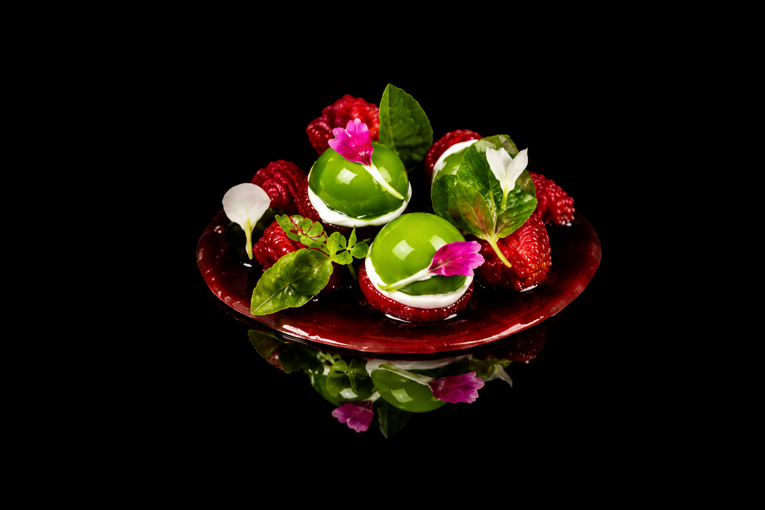 Dessert élégant avec deux sphères vertes garnies de fleurs roses, entouré de fruits rouges, de feuilles vertes et posé sur une assiette rouge brillant, avec une reflection sur une surface noire.
