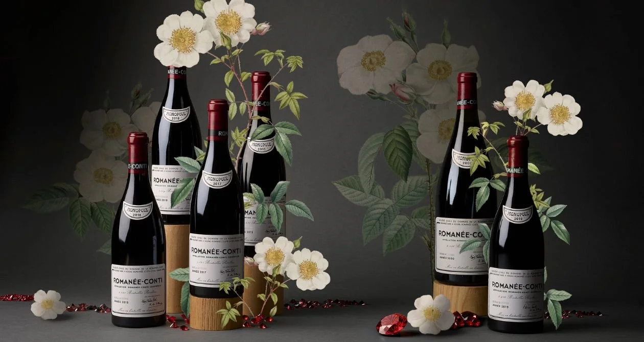 Cinq bouteilles de vin rouge romain, entourées de fleurs blanches et de feuilles vertes, disposées sur un fond sombre avec des pétales rouges.
