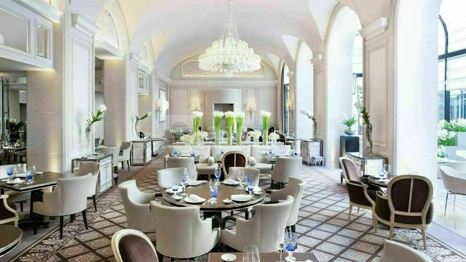 Salle à manger élégante avec des tables rondes, des chaises en velours et des décorations florales, dans un intérieur lumineux avec de grandes fenêtres et un chandelier au plafond.