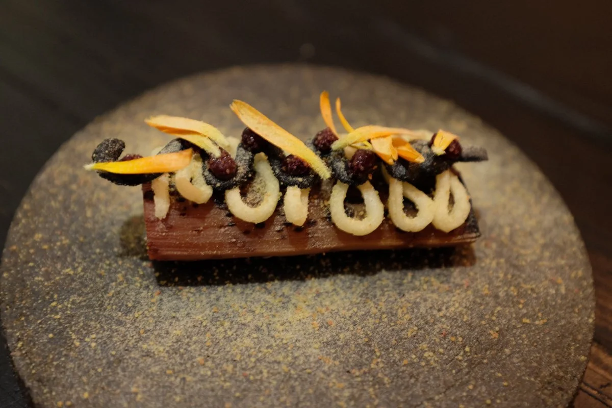 Un dessert chocolaté décoré avec des éclats d'amandes, des baies de genièvre et des articulations en chocolat, présenté sur une assiette noire