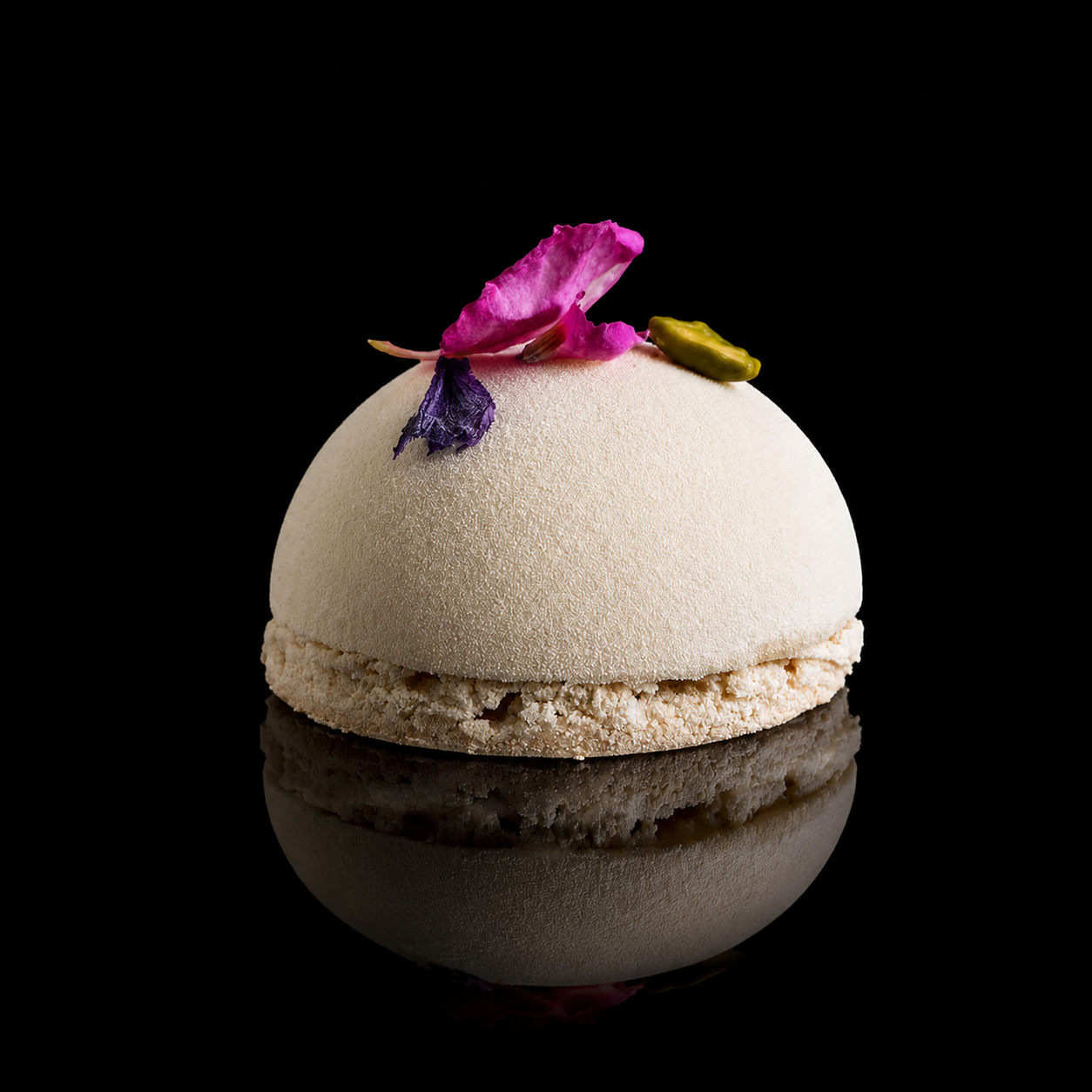Dessert rond blanc avec des fleurs séchées violettes, roses et un petit pistachier sur le dessus, posé sur une surface réfléchissante noire.
