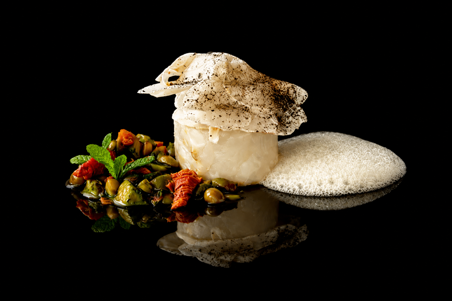 Plat gastronomique avec un fromage fondu sur un lit de légumes, accompagné de mousse et de garniture aux herbes, présenté sur une surface noire.