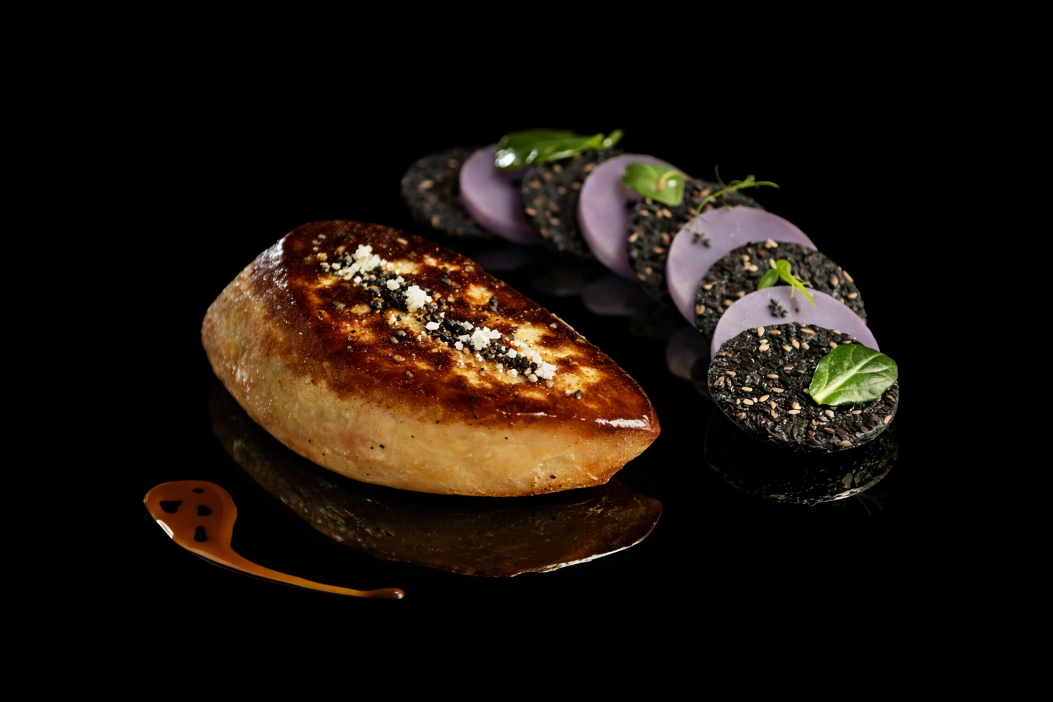 Assiette de cuisine gastronomique avec un morceau de foie gras laqué, une rangée de tranches de tache noire garnies de microgreens et de fleurs comestibles, et une petite décoration de sauce caramel sur fond noir.