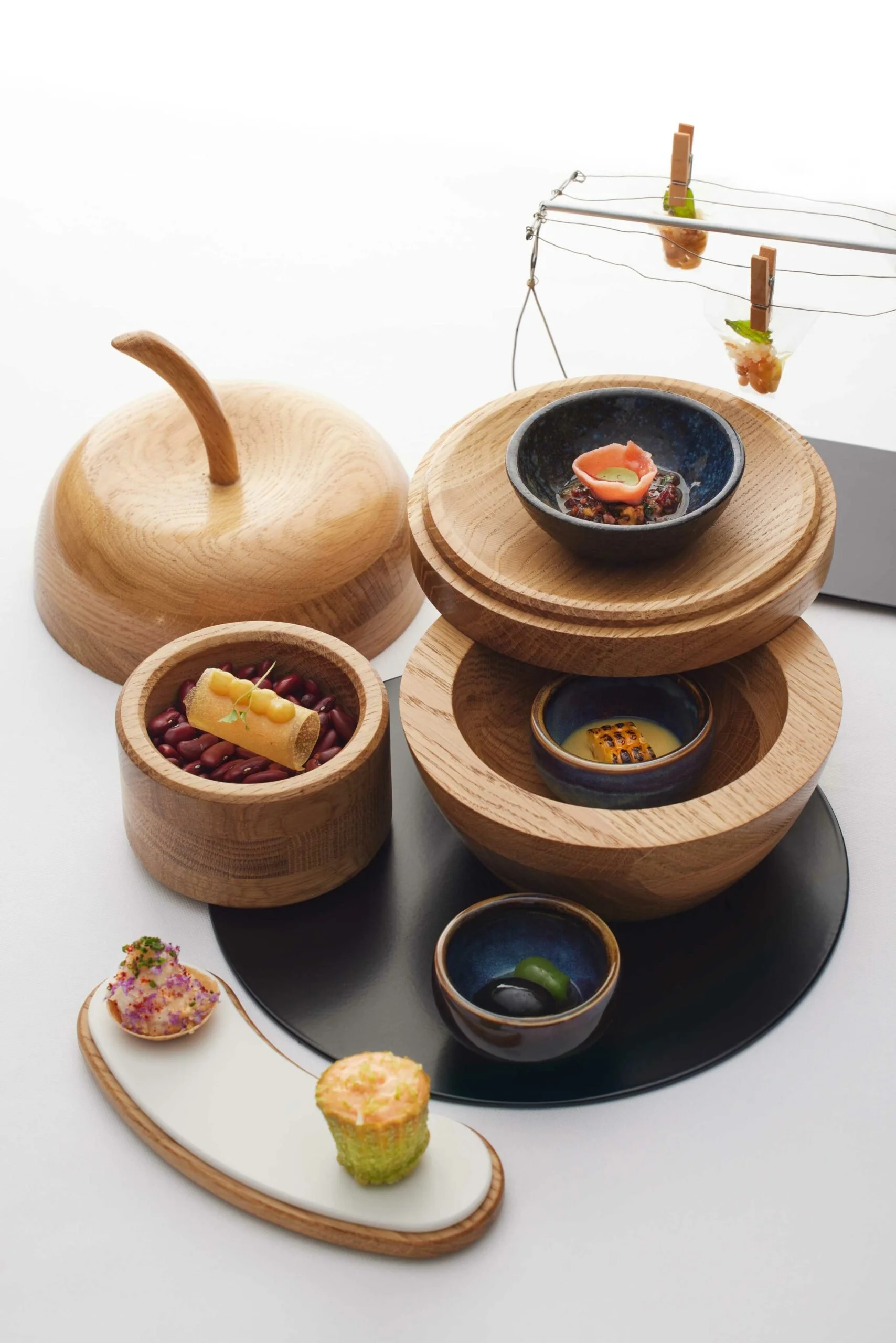 Présentation artistique de petits plats dans des bols en bois, avec des éléments décoratifs en bois et un plateau noir, comprenant des amuse-bouches et des amuse-gueules colorés.