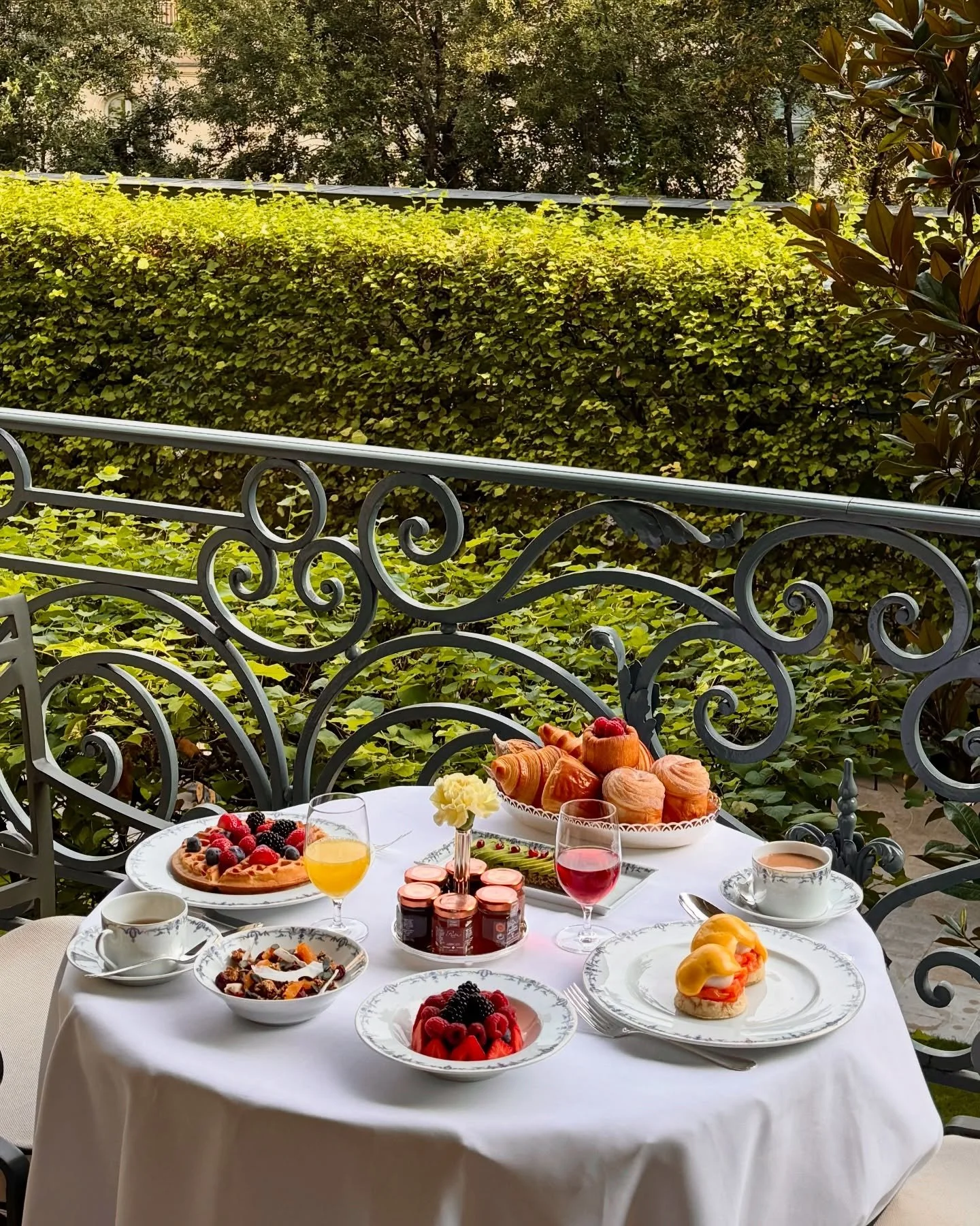Un petit-déjeuner ou brunch sur une table ronde avec des viennoiseries, des plats de fruits rouges, des boissons, un œuf à la coque, et une petite vase avec une fleur, en extérieur avec une balustrade en fer forgé et un jardin vert en arrière-plan.