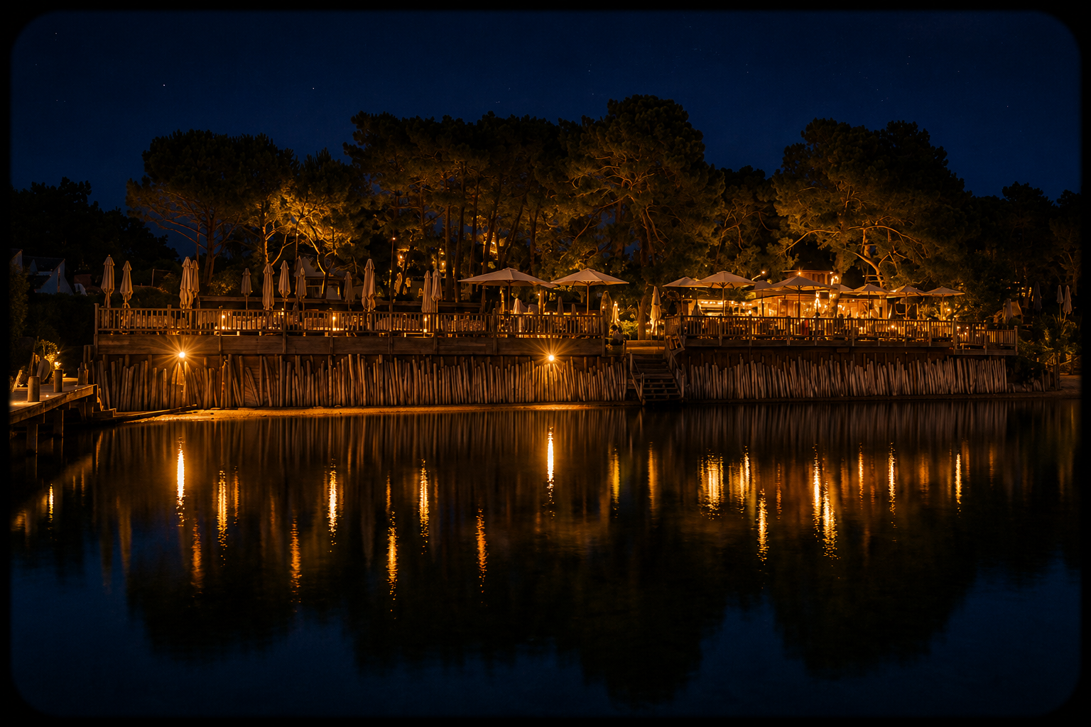 Terrasse d'un restaurant illuminée au bord de l'eau avec des parapluies, des arbres et des lumières douces en fond, la reflection dans l'eau.