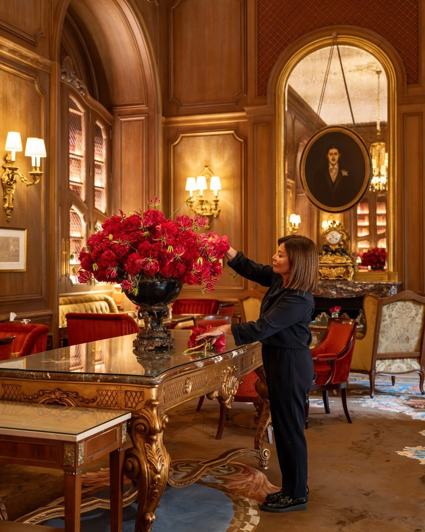 Femme décorant une grande vase de fleurs rouges dans un salon luxueux en bois avec des lampes éclairant la pièce.