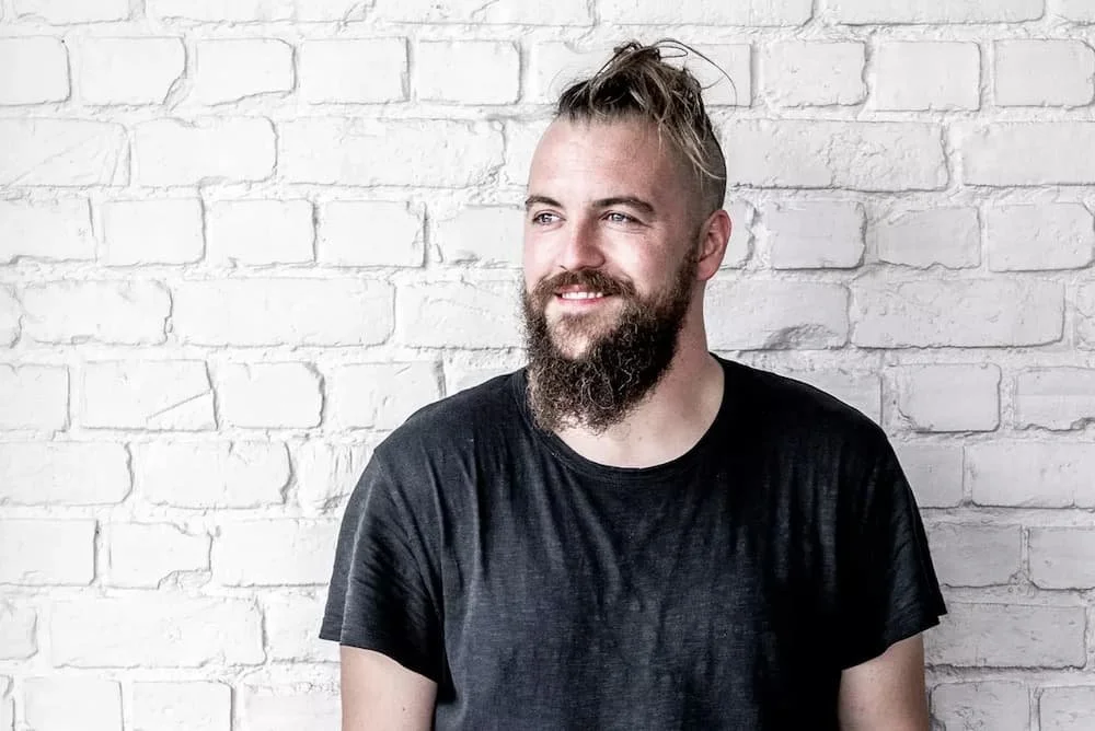 Un homme à la barbe et aux cheveux attachés, portant un t-shirt noir, debout devant un mur de briques blanches, souriant légèrement.