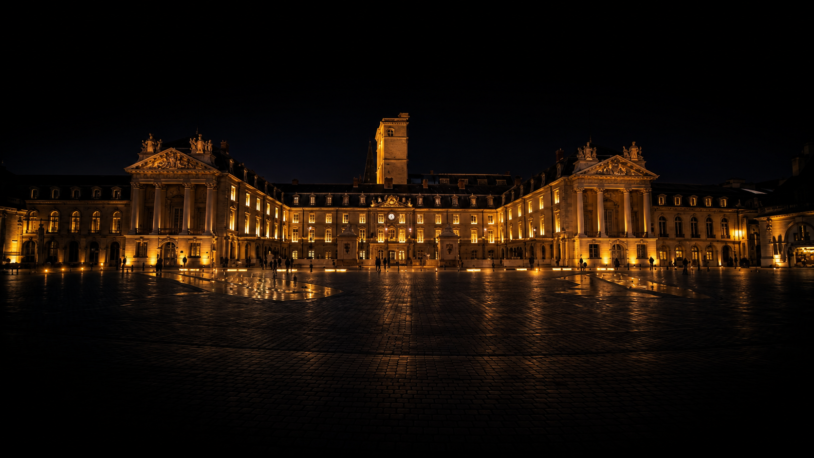 Bâtiment historique illuminé la nuit, avec une façade symétrique et des statues décoratives, reflété sur une place pavée humide.