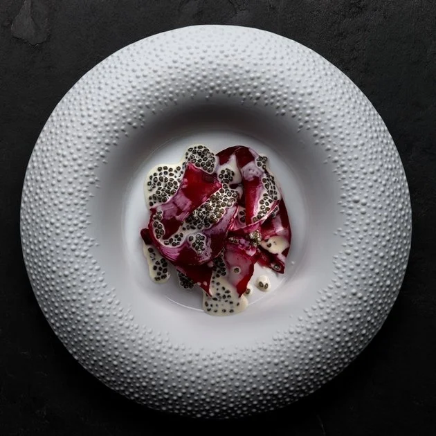 Dessert avec des tranches de fruit rouge, graines noires et blanc, servi dans une assiette blanche à texture ponctuée.