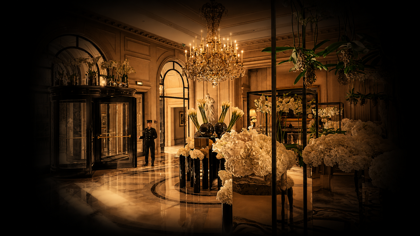 Un hall élégant avec un chandelier en cristal, des grands bouquets de fleurs blanches et une réception avec un portier en uniforme.