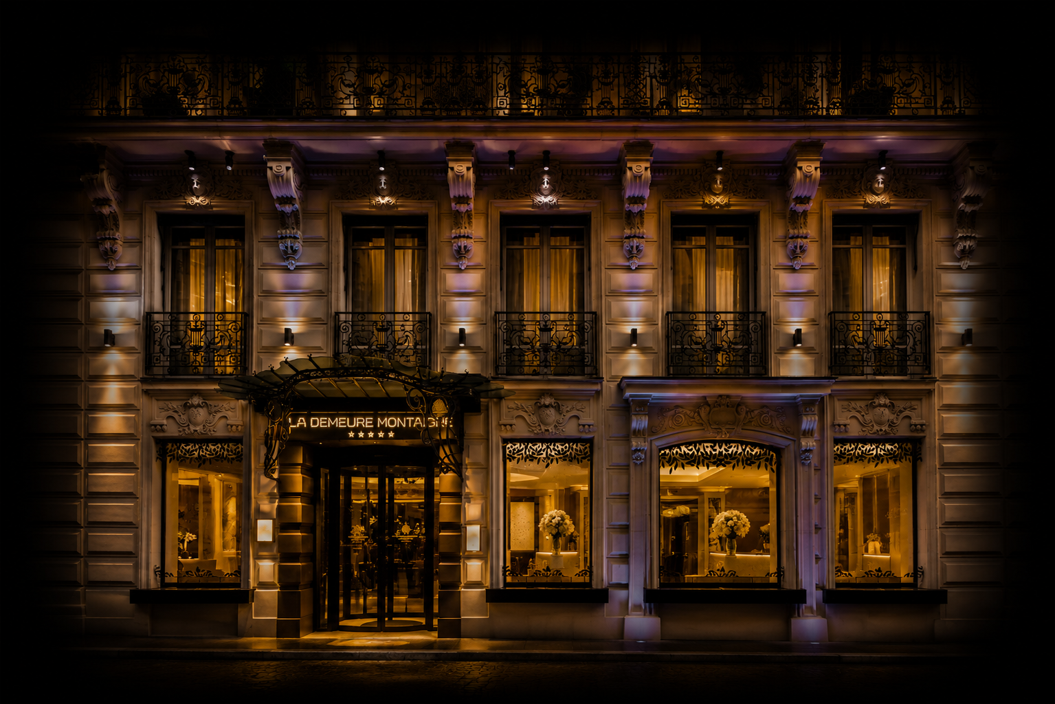Facade illuminée d'un hôtel avec enseigne 'La Demeure Montaigne', fenêtres avec rideaux et balcons en fer forgé, décoratif et éclairage chaud la nuit.