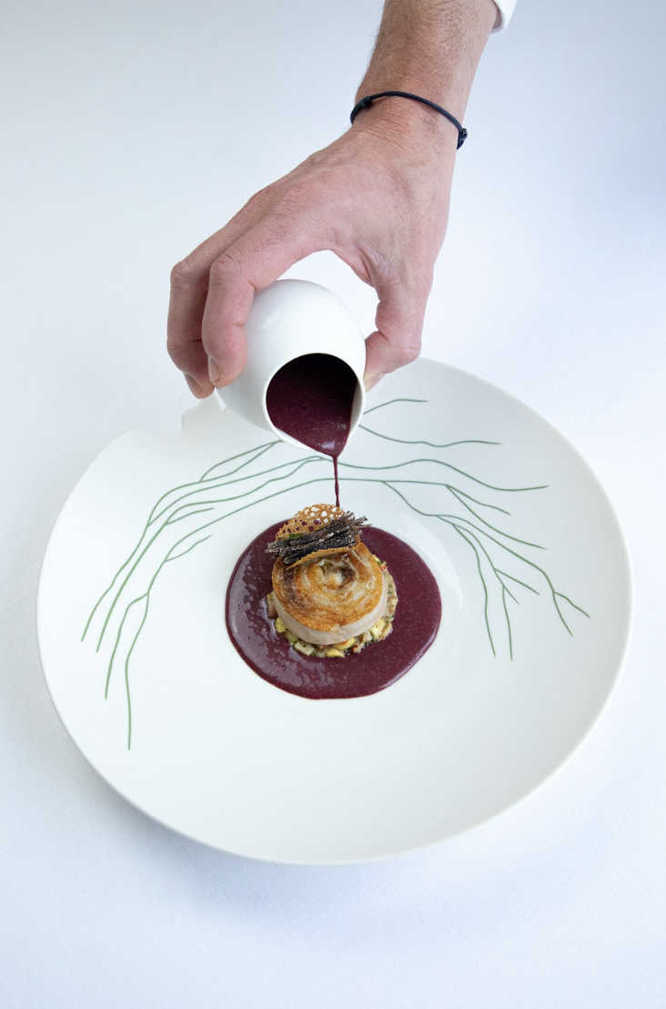 Une main verse une sauce bordeaux sur un plat gastronomique avec une garniture croustillante dans une assiette blanche décorée de lignes vertes