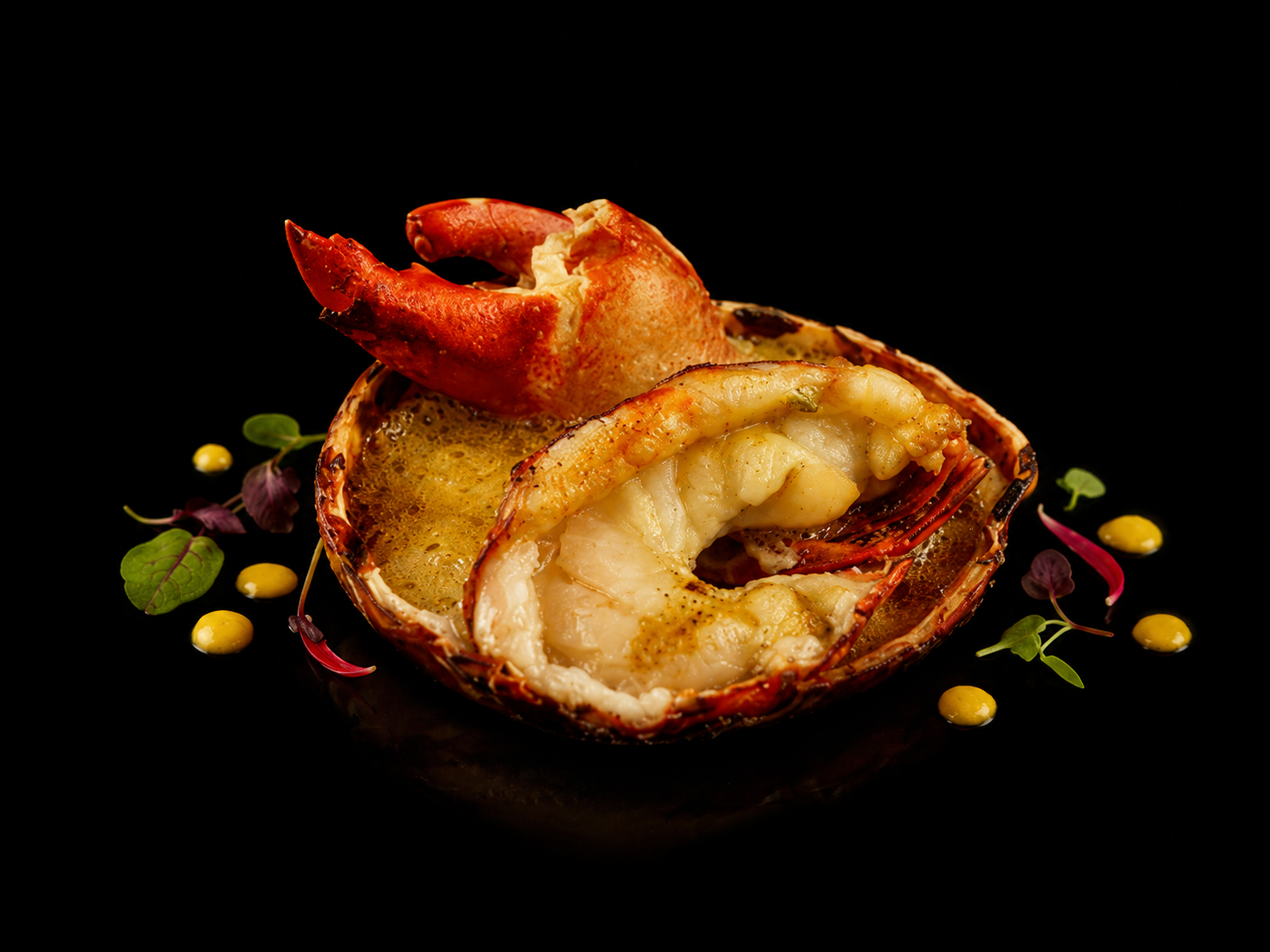 Gambas ou langoustines cuites dans leur coquille, présentées avec de la sauce et des mikros feuilles décoratives, sur un fond noir.