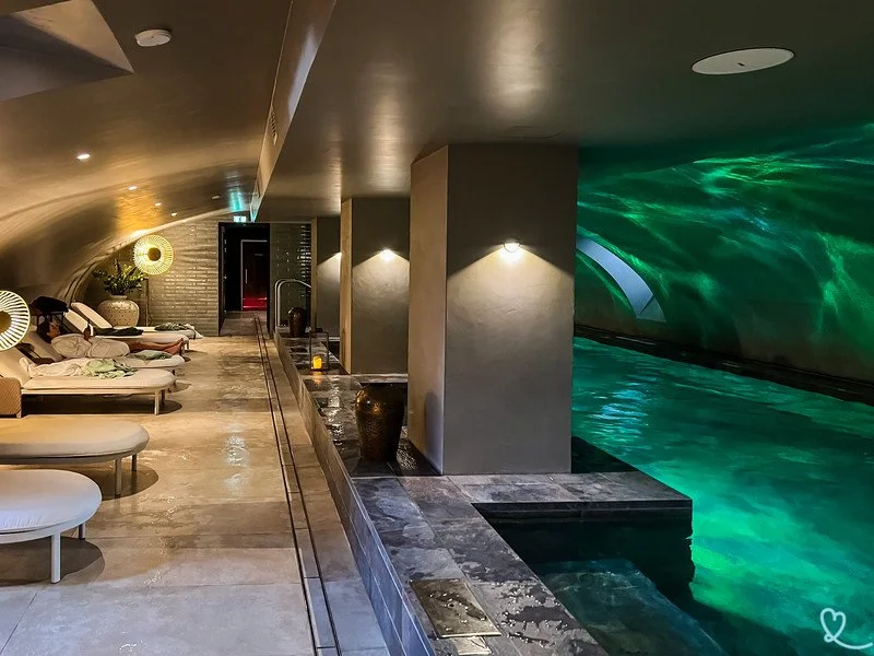 Piscine intérieure dans un spa ou un centre de bien-être, avec des zones de relaxation équipées de transats et de lampes de lecture, ambiance tamisée et atmosphère relaxante.