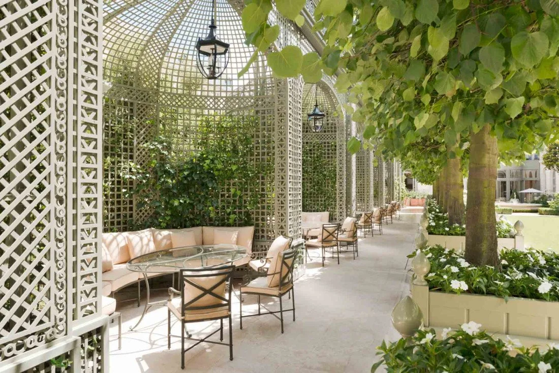 Terrasse avec pergolas en bois blanc, fauteuils avec coussins beige, plantes vertes, arbres, et lanternes suspendues.