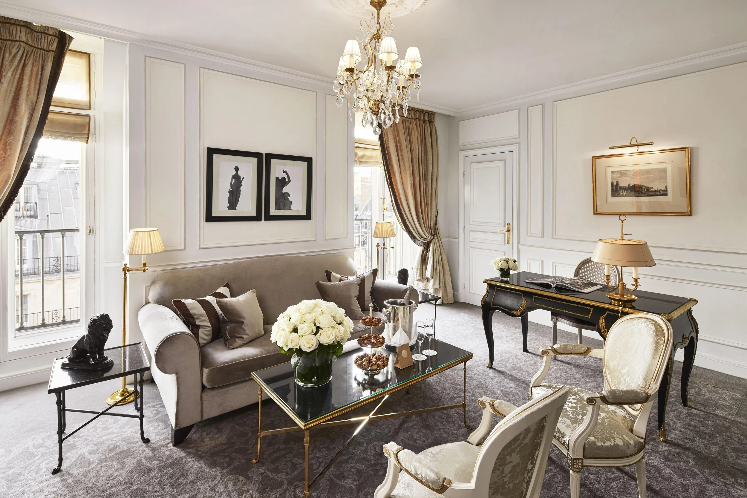 Salon élégant avec canapé gris, fauteuils blancs, table basse avec fleurs et collations, lampe, nappe, peinture encadrée, chandelier en cristal.