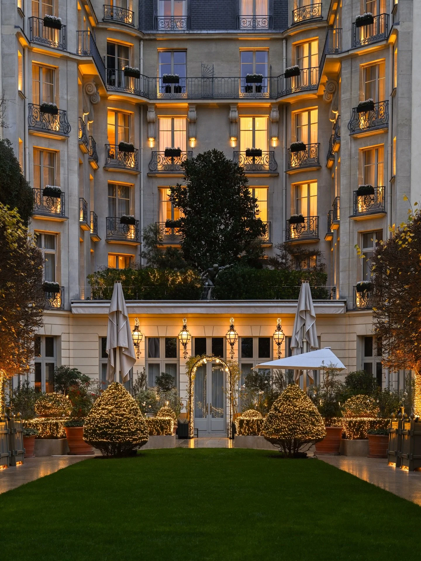 Vue d'un hôtel ou résidence avec plusieurs fenêtres éclairées à la nuit, une cour avec des arbustes décorés de lumières, un chemin, des parasols fermés et un portique décoré de guirlandes lumineuses.