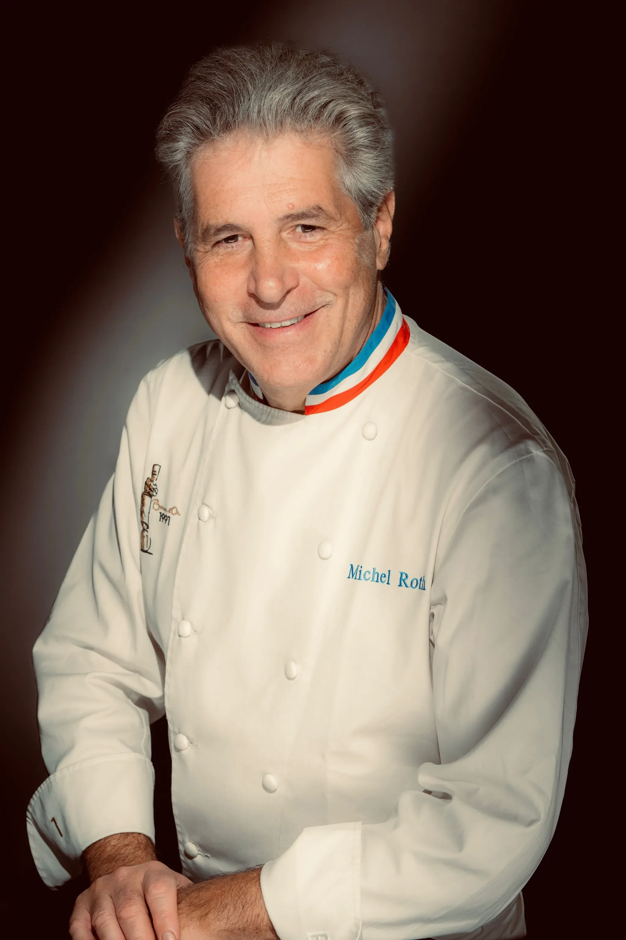Un chef en uniforme blanc, souriant, portant un tablier avec le nom "Michel Roth".