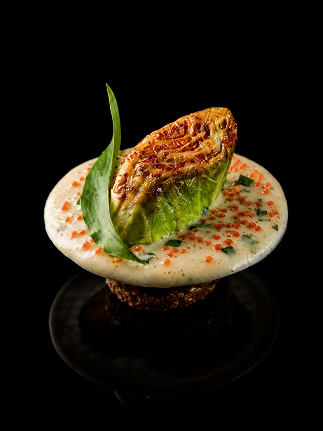 Une assiette conçue comme un plat de dégustation avec un artichaut grillé, entouré de sauce blanche, de petits grains rouges et de fines herbes, sur un fond noir.