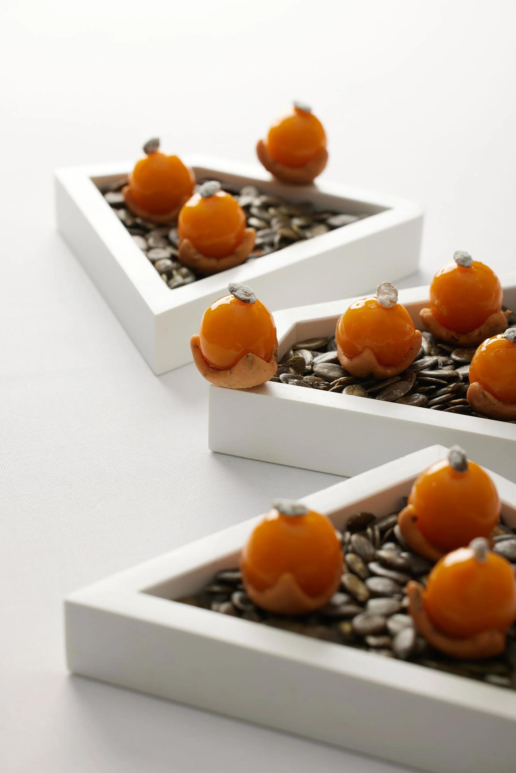 Gâteaux miniature en forme de fruits orange, décorés avec des petits éléments gris, présentés dans des paniers blancs avec des graines ou des céréales en dessous.