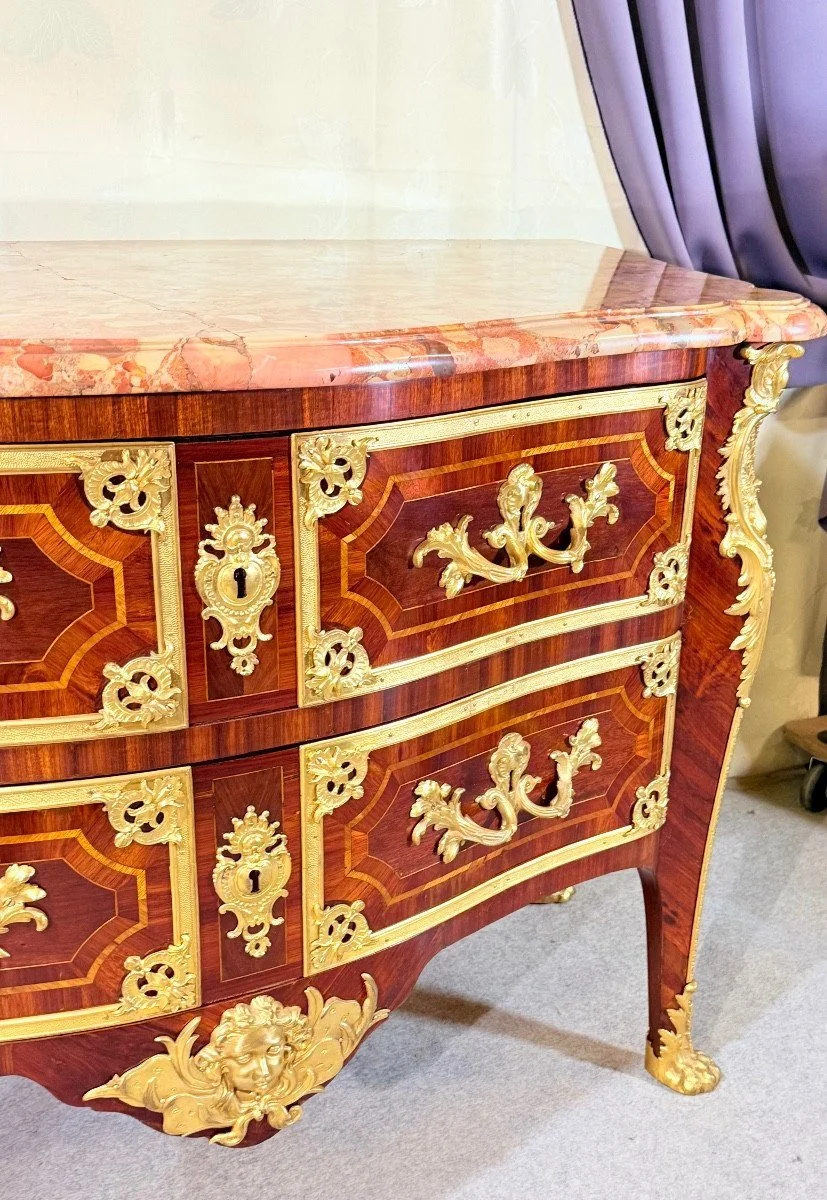 Un meuble ancien en bois avec des ornements en or, comprenant deux tiroirs, le dessus en marbre rose, et des détails décoratifs en or sur les coins et le bas.