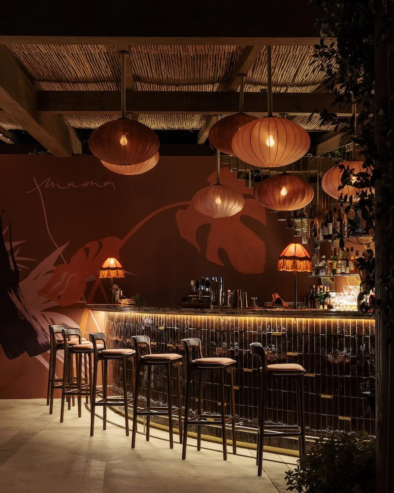 Un bar cosy avec un comptoir en carreaux noirs, des tabourets en bois avec des coussins, suspendu par des lampes rondes en papier ou tissu orange, ambiance chaleureuse et tamisée, décor floral mural et plantes vertes.