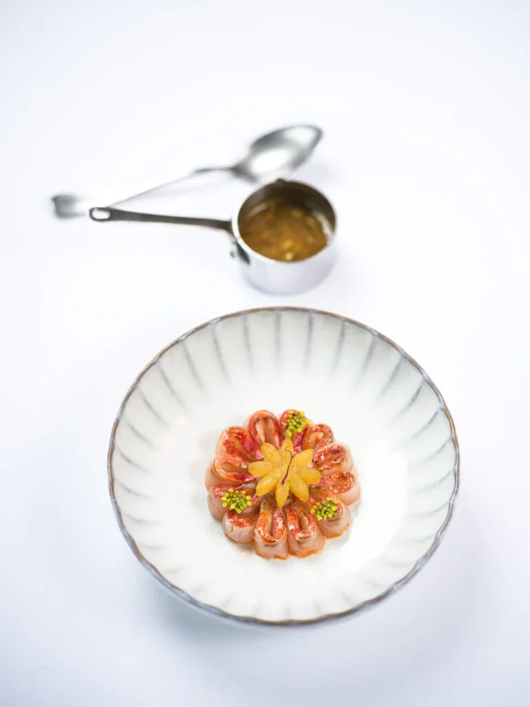 Une assiette blanche avec des crevettes garnies de petits morceaux de citron ou d'agrume, décorées de petites fleurs vertes, accompagnée d'une petite louche avec une sauce ou un condiment à côté.