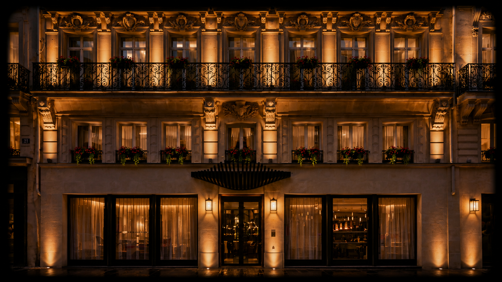 Façade illuminée d'un bâtiment européen avec balcons en fer forgé, fenêtres éclairées, décorations ornementales et fleurs sur les balcons, vue nocturne.