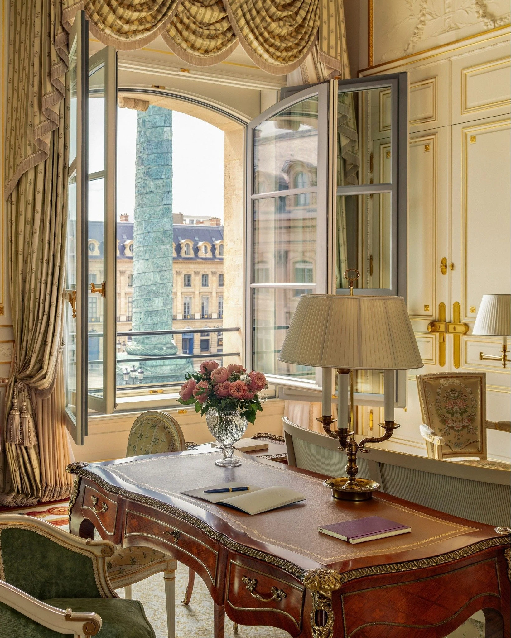 Une vue d'une pièce élégante avec un grand bureau en bois, une lampe à plusieurs branches, un vase avec des roses roses, des fauteuils et une grande fenêtre ouverte qui donne sur des bâtiments parisiens et une colonne en bronze.