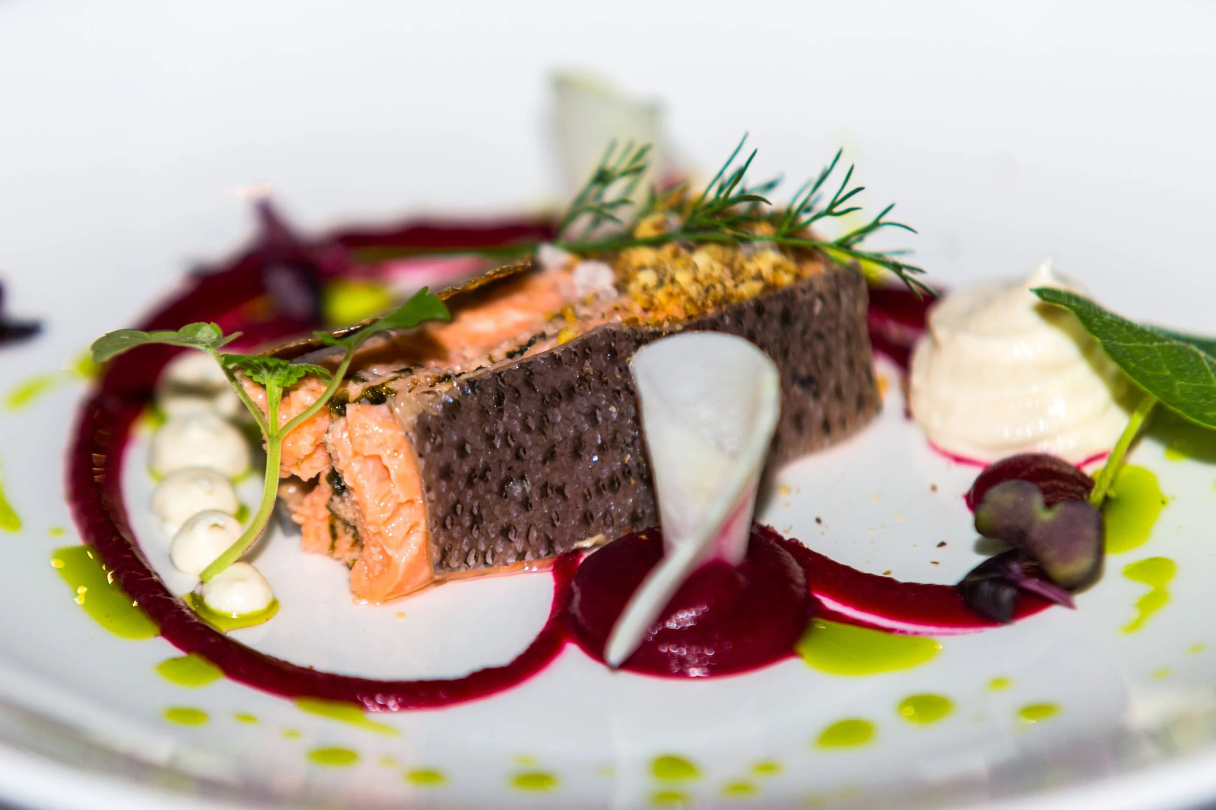 Plat gastronomique avec un morceau de poisson en croûte, servi avec des sauces et des herbes aromatiques, présenté de façon artistique.