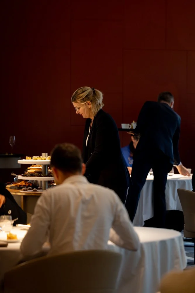 Une femme avec des cheveux blonds portant des lunettes, en tenue professionnelle, au buffet lors d'un événement ou dîner formel, avec plusieurs personnes en arrière-plan dans une salle de banquet ou réunion.