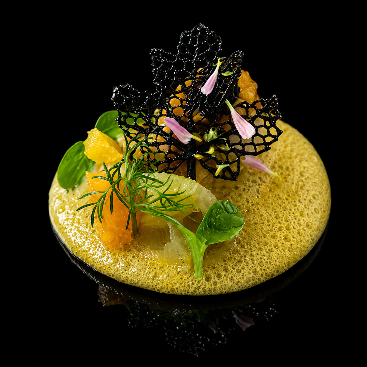 Une présentation gastronomique avec une mousse jaune, des feuilles vertes, des fleurs roses, un élément noir en forme de papillon, et des morceaux d'agrumes, sur fond noir.