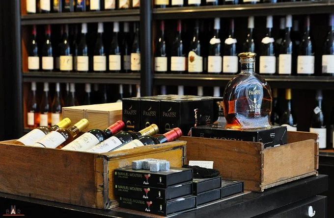 Vue d'une étagère avec plusieurs bouteilles de vin, un grand récipient en verre de cognac, et des boîtes de produits dans un magasin de vin ou de spiritueux.
