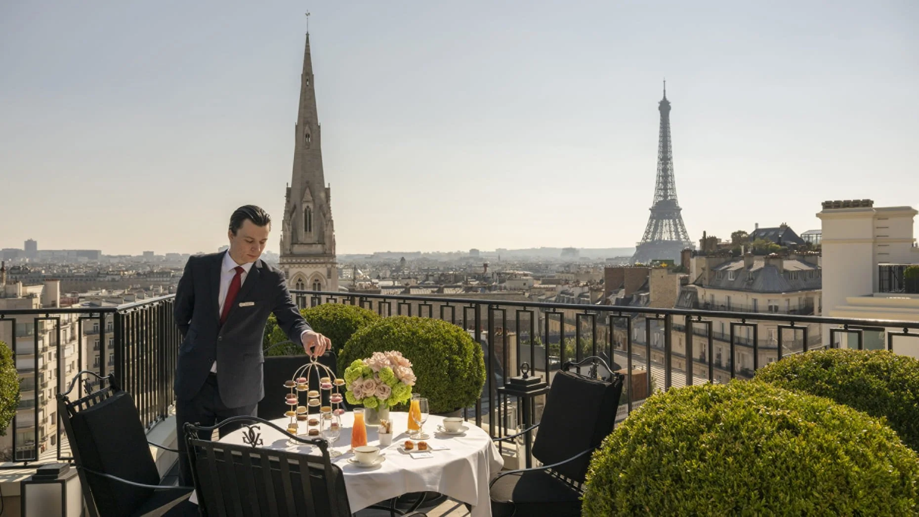 Un homme en costume noir avec une cravate rouge sert du thé ou du café lors d'un petit déjeuner ou goûter sur un balcon avec vue sur Paris, avec la tour Eiffel en arrière-plan.