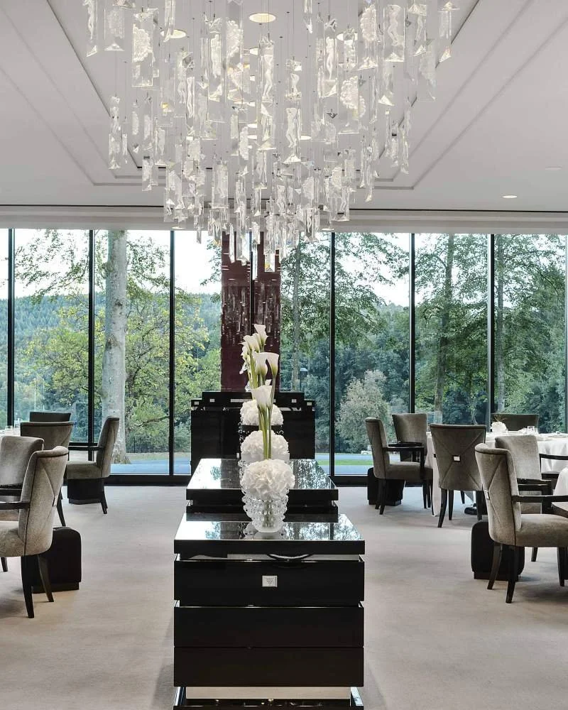 Intérieur d'un restaurant élégamment décoré avec un plafond en chandelier en cristal, grandes fenêtres avec vue sur la nature, tables et chaises en tissu gris, centre de table avec des fleurs blanches dans des vases.
