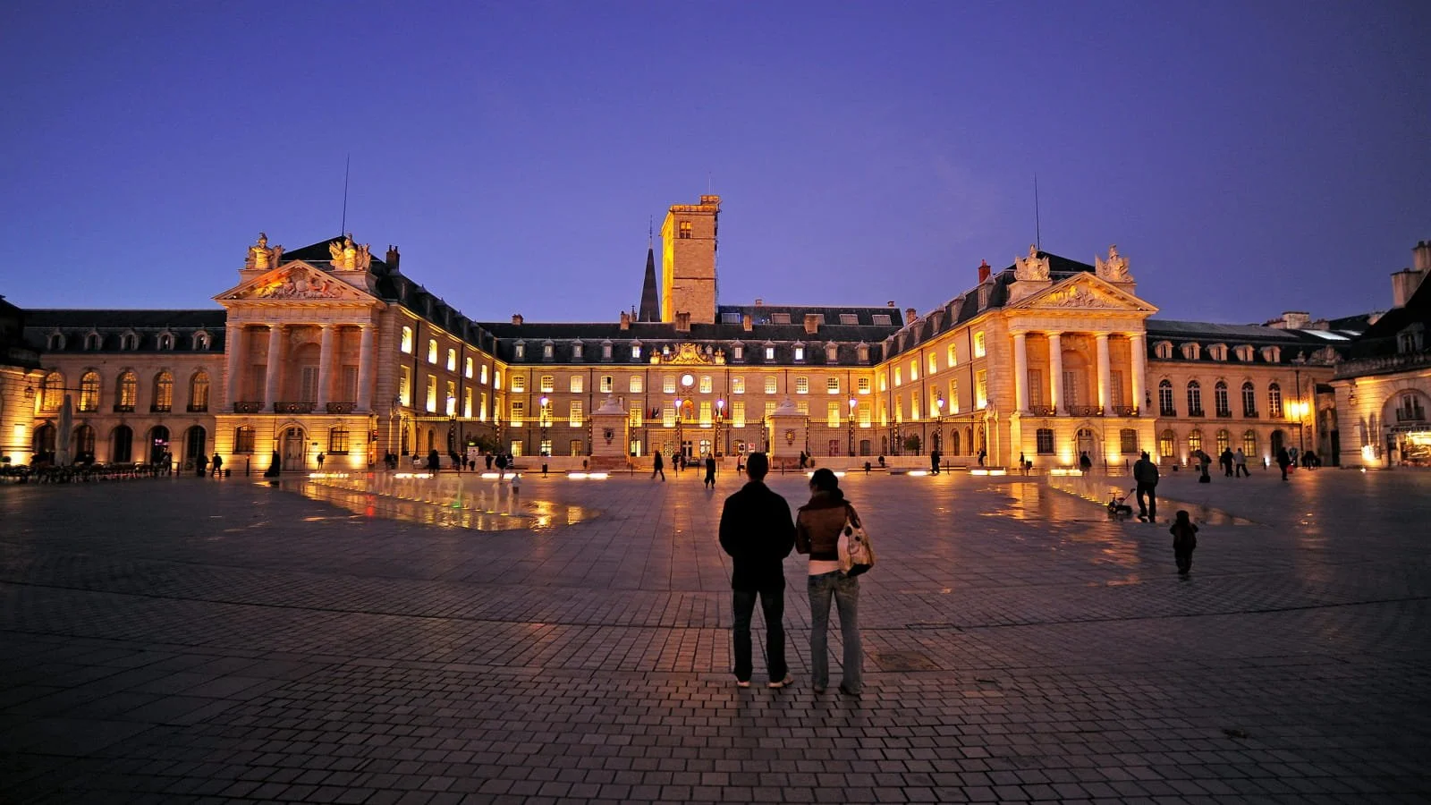 Une grande façade d'un bâtiment historique illuminé la nuit, avec des personnes qui se promènent dans la place pavée devant.