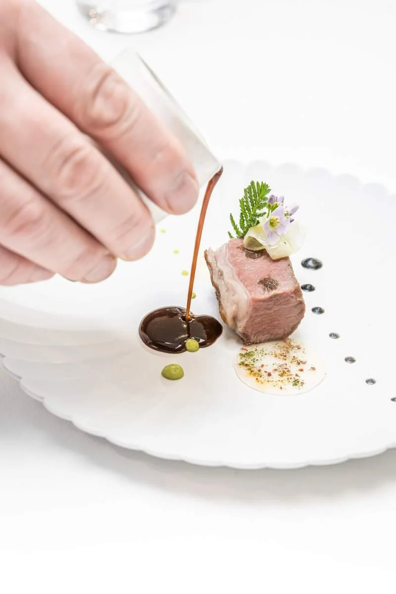 Un chef verse une sauce sur un plat gourmet avec un morceau de viande, décoré de fleurs et d'herbes, sur une assiette blanche.