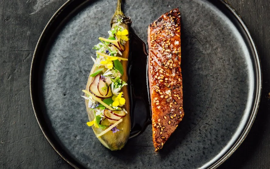 Un plat gourmet avec un poireau farci décoré de fleurs et une pièce de viande ou poisson glacée avec une sauce aux graines.
