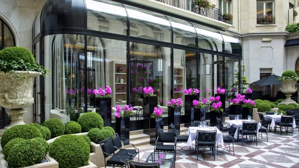 Terrasse de restaurant élégante avec tables, chaises, et décorations florales, entourée de bâtiments parisiens.
