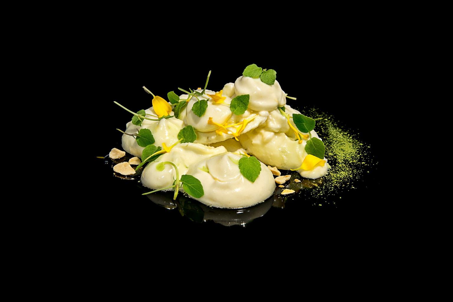 Dessert à base de glace ou de mousse blanche, décoré de feuilles vertes et de fleurs jaunes, avec des miettes et une poudre verte en accompagnement.